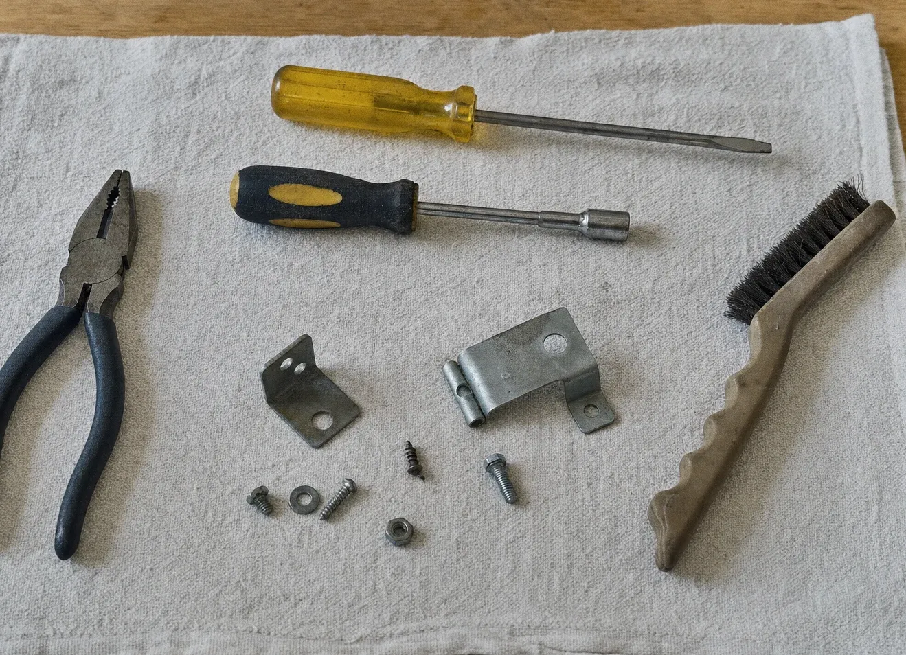 Pliers, screwdrivers, a nut driver and a wire brush laid out on a cloth with hinge brackets and loose screws